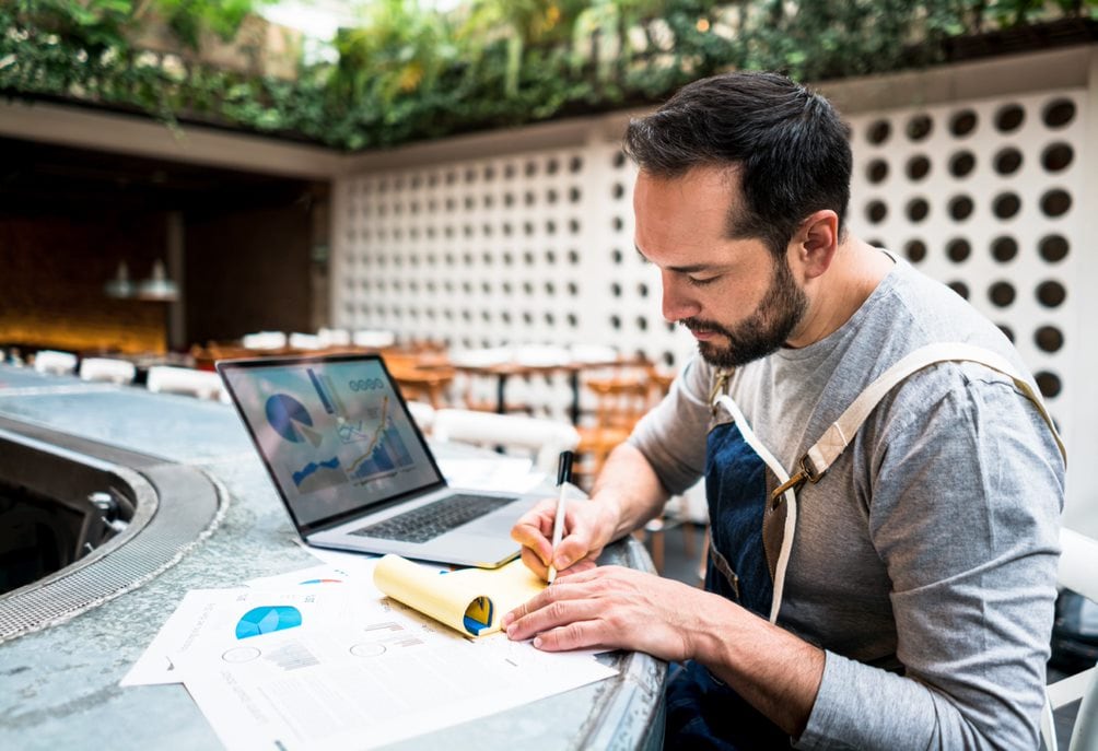 Man sitting at counter with laptop writing on notepad