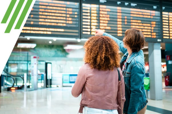 Two women at train station, one pointing at departure board