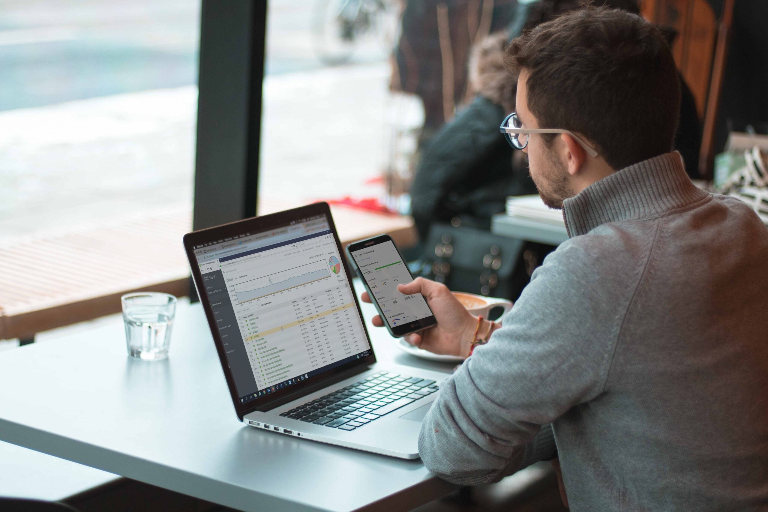 Man in a grey sweater sitting at a cafe wtih a laptop computer looking at his cell phone