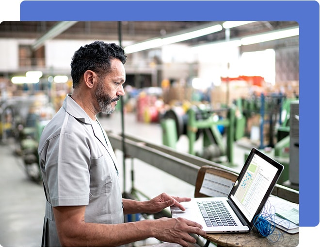 Man in a grey shirt standing in a manufacturing warehouse looking at a laptop computer